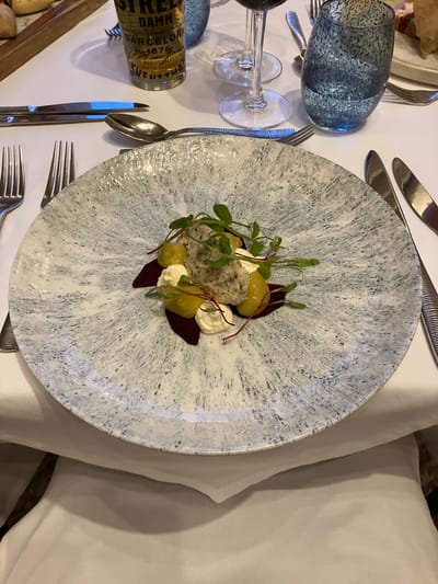 This is an overhead shot of a meticulously plated dish on a table, likely in a restaurant setting. The dish, centered on a blue and white speckled plate, consists of a mound of what appears to be shredded meat or fish, accompanied by spherical yellow items (possibly grapes or pickled vegetables), small dollops of white sauce, and slivers of a dark red ingredient, possibly beetroot. Microgreens provide a garnish.

The table setting includes silverware to the left and right of the plate. A blue-tinted drinking glass and a bottle of "Estrella Damm Barcelona" beer are visible on the left, along with a wine glass. The table is covered in a white tablecloth.

The scene appears to be indoors, in a formal dining environment. There's no specific indication of Glenridding, United Kingdom from the image itself.