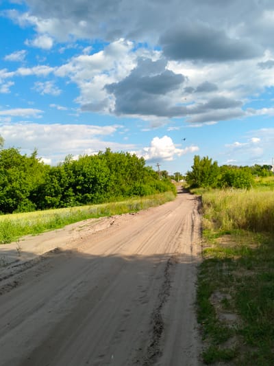 The image depicts a dirt road winding through a rural landscape in Matiashivka, Ukraine. The road is sandy with visible tire tracks, suggesting recent vehicle traffic. Lush green trees line the left side of the road, with a field of tall, golden grass to the right. In the distance, a few houses and utility poles are visible, hinting at a small village or settlement. The sky above is a vibrant blue, dotted with fluffy white and dark gray clouds, indicating partly cloudy weather. The lighting suggests it is daytime, likely late afternoon, as evidenced by the long shadows cast across the road. There is no visible text in the image. The scene evokes a sense of tranquility and openness, characteristic of a rural setting. The focus is on the natural landscape and the unpaved road, emphasizing a simple, country lifestyle.