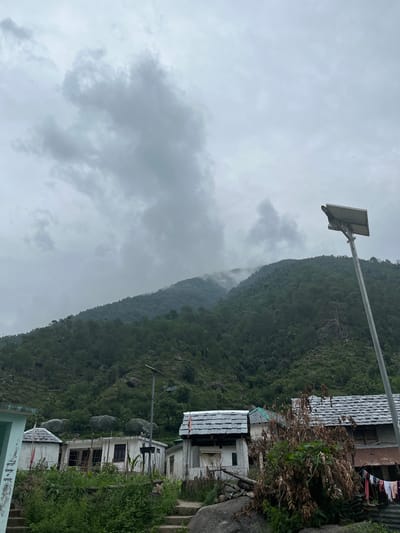 The image depicts a cloudy day in the city of Boh, India, with a focus on the natural landscape and local architecture. A steep, densely forested mountain dominates the background, with mist clinging to its higher slopes. In the foreground, a collection of small, white houses with grey slate roofs are clustered together. A solar-powered street lamp stands to the right of the houses, its panel angled towards the sky. Vegetation, including tall grasses and dry shrubs, surrounds the structures. Laundry hangs on a line in front of one of the houses. The overall scene suggests a rural or semi-rural environment in a mountainous region.
