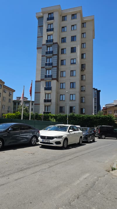 The image depicts a street scene in Ataşehir, Turkey, dominated by a tall, modern apartment building with a light-colored facade and dark-framed windows. The building has several floors, each with multiple windows and balconies. To the left of the main building, a portion of another, shorter building is visible, also with windows and balconies. Two flags, one white with a red symbol and another red with a white crescent and star, are flying on poles in front of the apartment building. In the foreground, several cars are parked or driving on the asphalt road. A dark-colored sedan is on the left, followed by a white Fiat sedan with the license plate "34 CAT 622". Behind the white car, a dark-colored SUV and another dark vehicle are partially visible. The sky is clear and blue, indicating a sunny day. A dense green hedge runs along the front of the apartment building, separating it from the road.