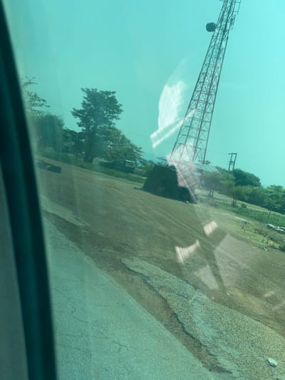 This media file depicts a scene in Jama'a, Nigeria, likely taken during the day, with overcast or hazy weather conditions indicated by the diffused light and the greenish-blue tint of the image, possibly due to reflections on a window.
The primary subjects include a large, red lattice metal structure, appearing to be part of a crane or a tower, that dominates the upper left portion of the frame. This structure is angled upwards and extends out of view. Below this, and to the right, is an area with a gravelly, dusty surface, resembling a construction site or an undeveloped plot of land. There are some trees and greenery visible in the background, suggesting a semi-rural or industrial setting. A dark, solid structure, possibly a building or a large piece of machinery, is situated near the base of the crane. Shadows are cast on the ground, indicating the presence of sunlight, though diffused.
The image seems to have been captured from a moving vehicle, as suggested by the perspective and the blurred foreground, which appears to be the interior of a car or truck. Reflections on what looks like a window obscure parts of the scene, creating a hazy and somewhat abstract quality. No people are clearly visible. The overall impression is one of an industrial or construction area, possibly undergoing development or operation. There is no visible text in the media file.