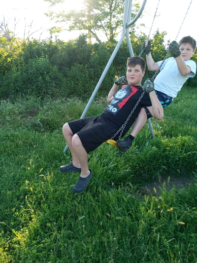 Two boys are on a swing set outdoors. The boy in the foreground is wearing a black t-shirt with a graphic of a face on it, black shorts, and black clogs. He is holding onto the swing chains with gloved hands. The boy behind him is wearing a white t-shirt, patterned shorts, and is also holding onto the swing chains. The swing set is made of grey metal. The background consists of green bushes and trees with sunlight shining through them. The ground is covered in green grass and some patches of dirt. The scene appears to be in a rural or suburban area, possibly a backyard or park. No specific landmarks or indications of the city of Zuya, Ukraine are visible in the image.