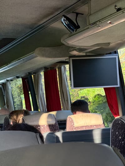 The image is a view from inside a bus, looking towards the front and right side. A flat-screen television is mounted on the ceiling in the middle of the frame, its screen dark. Below the television, several passengers are seated, their heads and backs visible. The seats appear to be upholstered with a patterned fabric and cream-colored headrests. Red and beige curtains hang near the windows, partially obscuring the view of trees and sunlight outside. The bus interior shows overhead compartments, lighting fixtures, and a rearview mirror. The overall lighting suggests it is daytime. There is no specific indication of the city of Kamburovo, Bulgaria, within the image itself.