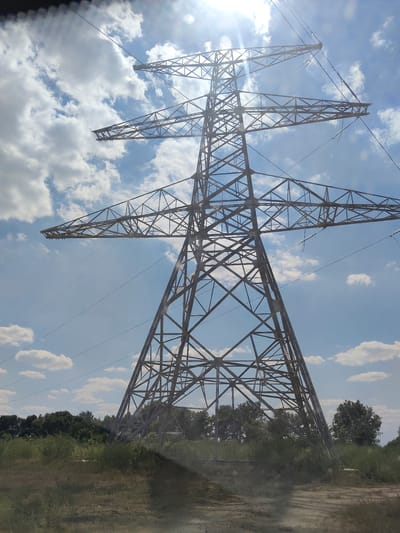 This media file captures a low-angle, wide shot of a large, lattice-steel power transmission tower against a bright, sunny sky filled with scattered white clouds. The sun is high in the sky, creating a lens flare and casting a strong light. The tower is the dominant subject, its intricate metal structure extending upwards and outwards, supporting thick electrical cables that disappear out of frame.

In the foreground, dry, unkempt grass and dirt suggest a somewhat rural or undeveloped landscape. Behind the tower, a line of dense green trees forms a natural backdrop. The overall setting appears to be an open area, possibly a field or undeveloped land, with the power line stretching across the terrain.

The weather is clearly sunny and appears to be daytime, likely late morning or early afternoon given the sun's position and intensity. There are no people or specific events depicted. The scene evokes a sense of scale and the power of infrastructure within a natural environment. There is no visible text in the image. The location context provided is Bilyaivka, Ukraine.