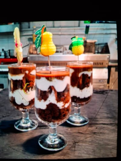Three layered desserts in clear glasses sit on a wooden surface. The desserts appear to be made of chocolate cake and white cream, with a reddish-brown topping. Each dessert is garnished with a decorative pick. One pick has a yellow, pear-shaped decoration with green leaves. Another pick has a yellow, pineapple-shaped decoration with green top leaves. The third pick is a pale yellow, possibly a flower or feather shape, with a pink accent. In the background, out-of-focus kitchen equipment and surfaces are visible. The image was taken at a restaurant in Medvezhyi Ozyora, Russia.