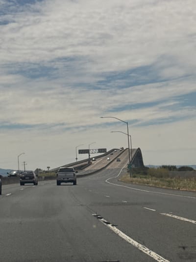 The image depicts a wide, multi-lane highway leading to a high bridge with a distinctive arch. The sky is partly cloudy, with white clouds spread across a blue expanse. The bridge appears to be concrete and steel, with traffic signs visible at its peak. Cars are traveling on the highway, including a silver pickup truck in the foreground and several other vehicles further ahead. The surrounding landscape consists of dry, grassy hills and sparse vegetation. The scene is captured from the perspective of someone in a vehicle on the highway, looking forward towards the bridge. The overall impression is of a vast, open highway infrastructure under a bright, partly cloudy sky. The location is identifiable as the Carquinez Bridge in Vallejo, California, United States of America, given its unique arch design and the surrounding topography.