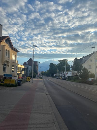 The image shows a street scene in Cottbus, Germany. The street is lined with buildings of varying heights and architectural styles. There is a wide street or roadway on the right, with tram tracks visible. On the left is a sidewalk constructed of pavers, with a red stripe running down the middle. There are streetlights and power lines overhead. Along the sidewalk, there are trash bins and signs, including one for Autohaus Blaskow, a local automobile repair shop, with telephone number 0355 42 77 77. The sky is mostly cloudy.