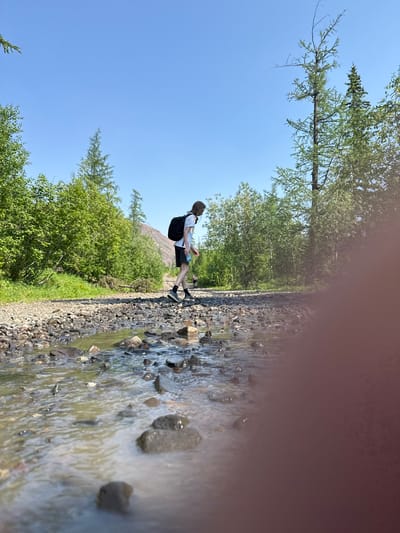 A young person with long, light brown hair, wearing a white t-shirt, black shorts, and a black backpack, walks across a shallow, rocky stream in a wooded area under a clear blue sky. The stream bed is composed of various sizes of grey and brown stones, with the water appearing slightly murky. Lush green trees line the path and the banks of the stream, with a brown, rocky mountain visible in the distance. The scene appears to be a hiking trail in the city of Talnakh, Russia, known for its natural landscapes. The image is taken from a low angle, close to the water's surface, and the foreground is blurred due to a person's hand being in the frame.