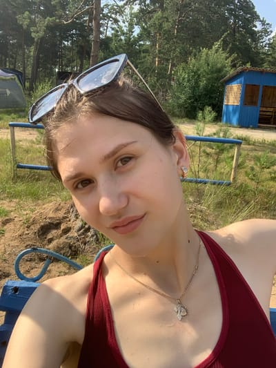 In a selfie taken outdoors, a young woman with light skin and brown hair is the primary subject. She is wearing a dark red ribbed top and sunglasses pushed up on her head. A thin gold chain with a pendant rests on her collarbone. The background suggests a rural or natural setting, with trees, dry grass, and a small blue wooden structure that appears to be a shed or cabin. The scene is illuminated by bright sunlight, indicating it's daytime, possibly midday or afternoon. The woman's face is turned towards the camera, and her expression is relaxed and pleasant, with a slight smile. The overall mood is casual and summery. The activity appears to be a relaxed moment outdoors, perhaps at a campsite or a recreational area, given the presence of the blue structure and what looks like part of a bench or seating area in the foreground. There is no visible text in the image.