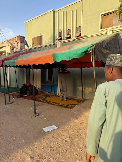 The scene depicts an outdoor setting in Daurawa, Nigeria, illuminated by bright daylight under a clear blue sky. A temporary, multi-colored canopy, segmented in orange, green, and black fabric, extends across the mid-ground, supported by dark metal poles.
Behind the canopy, a light-green, two-story building with multiple windows, some partially open, and vertical decorative moldings serves as a prominent backdrop. To its left, a more dilapidated building with exposed brick and two satellite dishes is visible. The foreground consists of dry, light-brown sandy ground exhibiting numerous footprint impressions and scattered debris, including white paper and clear plastic wrappers.
Under the canopy, four individuals are present. To the far left, two men are seated on a red and orange patterned rug. One wears dark clothing, and the other, also in dark clothing, wears a red cap. They appear to be engaged with an object or document on the rug between them. Centrally under the canopy, a third man stands barefoot on a yellow and black patterned prayer mat. He wears a light-colored tunic and has his hands raised towards his head, a gesture consistent with the beginning of Islamic prayer. In the immediate foreground, to the right, a fourth man is seen from the back, wearing a light green tunic and a patterned cap. His right arm is visible alongside his body.