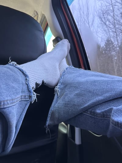 A pair of legs clad in distressed light blue denim jeans and white socks are elevated inside of a vehicle. The feet rest on the upper frame of what appears to be a hatchback or trunk opening. The interior features a dark, possibly leather, seat and a light-colored headliner. Through the vehicle's window, a snow-covered landscape with bare trees is visible. The image provides no indication of being in Dubrova, Russia.