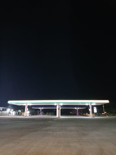 The image depicts a nighttime scene at a Petrobras gas station in Pailitas, Colombia. A wide, illuminated canopy covers multiple gas pumps. The ground is paved with concrete, and in the background, there are some low buildings and trees. The sky is dark and clear. Two figures are seated on a yellow bench under the canopy, appearing to be resting or waiting. The scene is generally quiet and appears to be unoccupied by other customers.