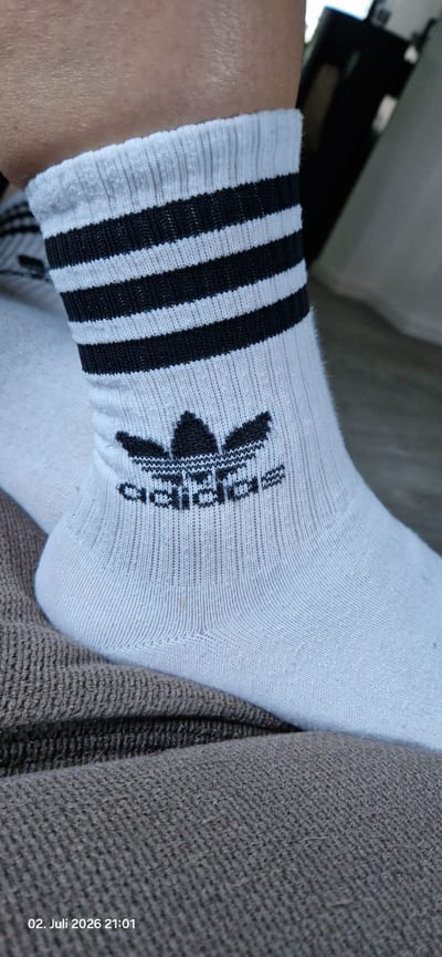 A close-up, low-angle, full shot shows a person's right foot wearing a white ankle sock with black Adidas branding and three black stripes near the cuff. The sock is positioned on a brown, textured surface, possibly a couch or blanket. The background is blurred, showing a dark, indistinct object on the right and a lighter surface on the left. A timestamp in the lower-left corner reads "02. Juli 2026 21:01". The image does not provide enough information to determine the specific location within Hummeltal, Germany.
