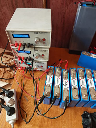 The image depicts a close-up, high-angle view of a wooden table with various electronic components and equipment, likely in a workshop or storage area. The primary focus is on several blue rectangular battery cells arranged in a row. These cells have silver terminals and white labels with barcodes and symbols. Some of these cells are connected to a set of battery testers or electronic load devices stacked vertically. The testers have digital displays showing voltage and amperage readings, and are connected by red and black wires with alligator clips. A white power strip with three plugs is visible on the left side, connected by a black power cord. In the background, a portion of a large, dark gray rectangular object, possibly another battery or a power supply, is visible. A framed image of Che Guevara is also present on the table. The overall impression is one of a DIY or hobbyist electronics setup. There is no identifiable information within the image suggesting a specific location in Cheremkhovo, Russia.