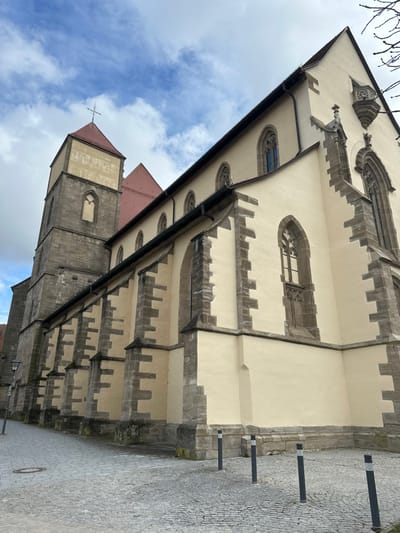An exterior, low-angle photograph depicts a prominent historic church building situated on a cobbled street in Weißenburg in Bayern, Germany. The structure features a main body with pale yellow plastered walls and robust dark grey stone buttresses and window surrounds. Multiple tall, arched windows with intricate gothic tracery are visible along the side, some containing dark panes. A dark rain gutter system runs along the eaves beneath a steeply pitched red-tiled roof. To the left, a square-section stone bell tower rises, topped with a red pyramidal roof and a simple metal cross. This tower features a single arched window opening near its apex. The far right end of the building displays a more elaborate gothic-style window and a small, decorative stone balcony or niche high on the gable. The foreground consists of a grey cobblestone pathway, with a circular metal manhole cover positioned towards the lower left. Four dark bollards, each with a white reflective band, are arranged in a linear pattern on the cobblestone near the church's base on the right side. The sky above is partially cloudy, revealing patches of blue. No individuals, vehicles, or discernible actions are present in the scene.
