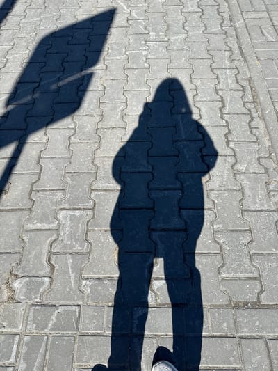 The image shows a shadow cast on interlocking gray pavement stones. The shadow appears to be that of a person wearing a hat, with the shadow of their legs extending to the right. A portion of a person's shoe, likely connected to the legs, is visible in the upper right. The location is implied to be the city of Beloyarsky, Russia.