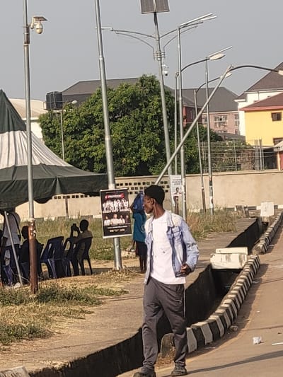 The image depicts an outdoor urban scene in San Martín, Colombia, under clear daylight. In the immediate foreground, a Black male, dressed in a white t-shirt, light blue denim jacket, dark grey sweatpants, and black clogs, walks towards the right of the frame on a paved path, looking to his left. To his left, a concrete drainage ditch with black and white striped markings runs parallel to the path.
In the mid-ground, to the left, a large tent with alternating dark green and white stripes is set up on a grassy area. Underneath the tent, several blue plastic chairs are arranged, some occupied by indistinct figures. One figure is clearly visible seated, holding what appears to be a mobile phone to record or photograph. Adjacent to the tent, a black poster displaying "PRECISION 5" and "CEZA CITY MUSIC," along with images of multiple individuals, is affixed to a metal pole. Behind this pole, a partially obscured person wearing a blue head covering stands.
Multiple grey metal poles are distributed throughout the scene, supporting streetlights with various designs, some featuring solar panels and integrated dome-shaped CCTV cameras. In the background, a large green tree is prominent, flanked by several buildings with different architectural styles, including a distinct yellow building on the far right. The ground consists of paved areas and unkempt grassy verges.