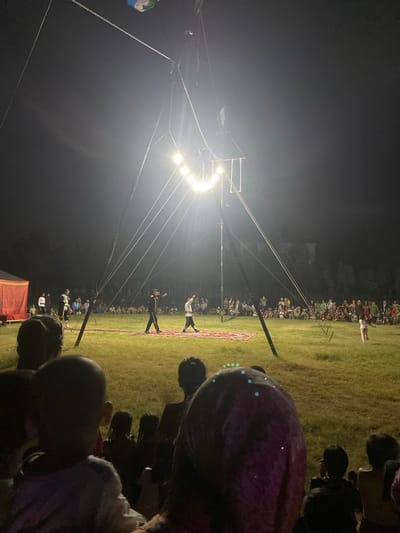 An outdoor circus performance is underway at night in Arbagish, Uzbekistan, illuminated by bright stage lights strung from a tall, tripod-like structure. The performance area is a grassy field with a red carpet laid out for the performers. Three individuals are visible on the carpet, dressed in performance attire. One, in the center, appears to be leading the performance, gesturing with his arms. To his left and right, two other performers are also engaged in the act, one of whom is walking with a raised hand. A large audience, comprising adults and children, is seated on the grass, watching the show. In the background, a tent with a red curtain is set up on the left side of the field. The overall scene depicts a community gathering for an evening of entertainment in a rural or park-like setting within Arbagish.