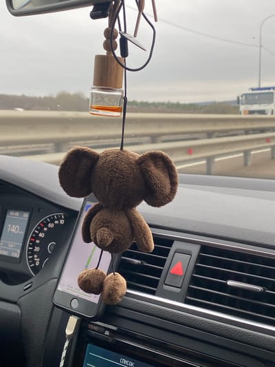 The image is taken from inside a car moving on a highway, likely in Russia given the language on the dashboard display. A brown plush toy (likely a bear) hangs from the dashboard, partially obscuring a smartphone that is mounted on a phone holder. The smartphone displays a map. An air freshener hangs from the rearview mirror. The car's speedometer is visible and indicates a speed of 140 km/h. The dashboard also features air vents, an emergency flasher button, and a built-in entertainment system screen. The highway outside is multi-laned, and the weather appears overcast. The city of Novyy Shardak is not discernable in this image.