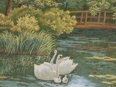 This is a still life painting of a serene natural landscape. The scene depicts a tranquil body of water, likely a pond or lake, with lush green reeds and bushes lining the banks. A wooden bridge arches gracefully over a section of the water in the background. In the foreground, two adult swans and two cygnets are swimming peacefully. The swans are depicted in a classic white, their elegant necks curved. The water reflects the surrounding greenery and the bridge, creating a harmonious and picturesque scene. The painting style is impressionistic, with soft brushstrokes and a gentle color palette. The specific location in the city of Motmos, Russia, is not identifiable from the image itself, as it presents a generic natural setting.