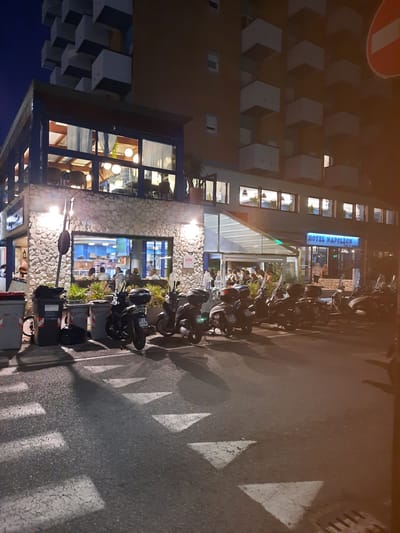The image depicts the exterior of the Hotel Napoleon in Pesaro, Italy, at night. The hotel has a modern architectural style with multiple floors, featuring numerous rectangular balconies. At the street level, there is a restaurant or cafe with a stone-clad facade and large windows, revealing patrons inside. A covered outdoor seating area is also visible, with people gathered.

Parked along the street in front of the establishment are several scooters and motorcycles. The road in the foreground features a pedestrian crossing with white triangular markings. Trash bins are placed along the sidewalk. The lighting suggests it is nighttime, with streetlights and interior lights illuminating the scene. The sky is dark blue, indicative of the evening or night.