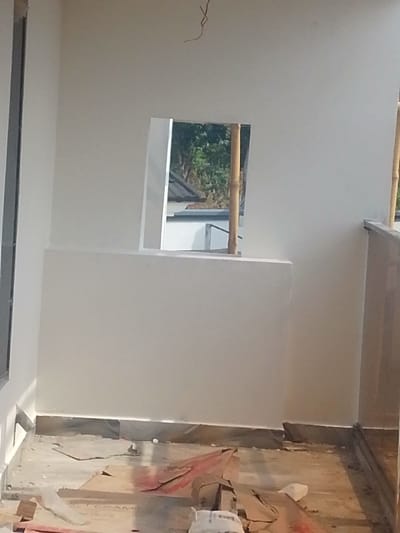 An interior view depicts a room or balcony in Agukwu Nri, Nigeria, undergoing construction or renovation. The space features freshly painted white walls, with a dark gray trim lining the base. A rectangular opening in the center of the far wall acts as a window, revealing an exterior scene with distant green foliage, a dark-tiled roof of an adjacent building, and a lighter structure below it. A bamboo pole is visible vertically through the right side of this opening, possibly part of external scaffolding. The concrete floor is scattered with construction debris, including crumpled cardboard pieces, one of which bears the partial label "VICTORIA TILES." Shadows are cast onto the floor from the left. A dark gray vertical frame, possibly for a door or window, is visible on the far left wall. A low wall or railing with a dark top edge is partially visible on the far right, suggesting a balcony edge. A thin, dark, curly object hangs from the white ceiling near the top center of the frame. No individuals, explicit actions, or interactions are present.