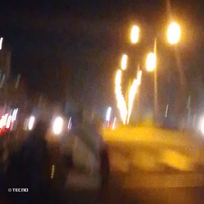 A blurred nighttime scene with bright lights dominating the view. Two prominent streetlights illuminate the area. Streaks of light suggest movement, possibly from vehicles. A group of people can be partially seen in the lower left, their forms indistinct due to the motion blur. The overall impression is of a busy street at night captured with a slow shutter speed. A 'Tecno' watermark is present in the bottom left corner.