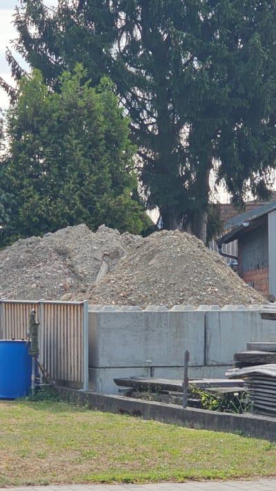 A construction site in Vöhringen, Germany, shows large piles of rubble and dirt in the background, against a backdrop of mature evergreen trees and a building. In the foreground, there is a blue barrel next to a corrugated metal fence and a hand pump. Behind these, concrete blocks are stacked, and further to the right, there are stacks of wooden planks. The ground is covered with grass and a paved walkway. The scene appears to be a storage or staging area for construction materials and debris.
