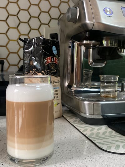 The image depicts an indoor kitchen environment in Ridgefield, United States. A clear glass containing a layered beverage, appearing to be a latte with distinct white foam, light brown, and white layers, is positioned in the foreground on a light-colored, speckled countertop. Behind the glass, two bags of ground coffee are visible; the prominent bag is black with "BAILEYS Irish Cream" branding and "French Vanilla" text. To the right, a silver-toned espresso machine, resembling a Breville model, rests on the countertop with a glowing purple "POWER" button illuminated. A portafilter is attached to the machine, and two small, clear glass vessels with coffee residue are on its drip tray. A light-colored mat with a green leaf pattern is under the espresso machine. The background features a backsplash composed of hexagonal tiles with golden or bronze outlines and a white electrical outlet. No individuals are present in the scene.