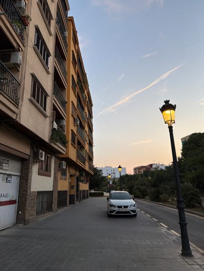 The image depicts a street scene in Benimàmet-beniferri, Spain, during what appears to be sunset or early evening. A white car, a Seat Leon with license plate 6389 KZD, is parked on the right side of the street, facing forward. The car is positioned on the asphalt roadway, next to a sidewalk paved with grey rectangular tiles.
On the left side of the street, a multi-story building with beige and yellow facades dominates the view. The building has balconies with black railings, and windows with dark shutters. Air conditioning units are visible on the exterior walls. A large garage door with red and white stripes is partially visible at the bottom left of the frame, with a "SE ALQUILA" (for rent) sign.
The street is illuminated by a vintage-style black lamppost with a glowing yellow light, positioned on the right side of the sidewalk. In the background, several other lampposts are visible, along with trees and some residential buildings. The sky above is a gradient of pale blue to orange, with faint clouds and contrails from an aircraft. The overall impression is a quiet urban street at dusk.