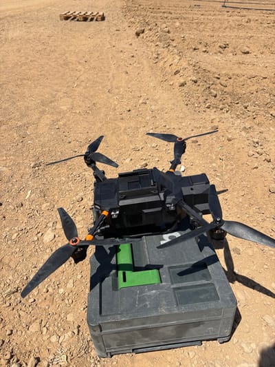 A black quadcopter drone with four propellers and orange structural accents rests on a dark gray modular storage case. The case has a bright green rectangular insert on its lid. The scene is an open, unpaved field characterized by light brown dirt and small rocks. Several wooden pallets are visible on the ground in the background. The environment is illuminated by direct sunlight, casting shadows from the drone and case. No individuals are present. The location is an outdoor, uncultivated area within the Merhavim Regional Council, Israel.