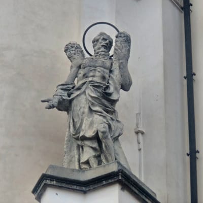 A low-angle, medium shot captures a weathered, stone statue of a winged figure with a halo, mounted on a building in Calcinate, Italy. The statue depicts a male figure with muscular arms, clad in draped clothing, with both hands raised and holding what appear to be scrolls or bundles. The figure's face is weathered and somewhat obscured by the halo, which is a thin, dark circle. The statue is positioned in front of a light-colored, textured building facade. A dark, vertical metal pipe is visible on the right side of the image, attached to the building. The statue rests on a decorative stone base.