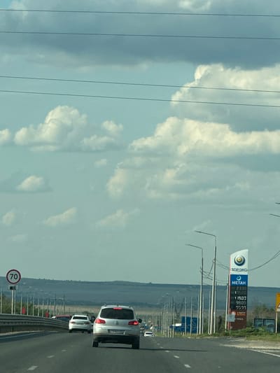 The image shows a highway scene in Zorinskii, Russia. The view is from a vehicle traveling on a multi-lane asphalt road. On the right side of the road, there is a gas station with a prominent sign displaying fuel prices, including "METAH" priced at 23.90, 19.00, 98.00, and 29.97. Several streetlights line the right side of the highway. On the left side of the road, there is a speed limit sign indicating "70". Several cars are visible on the road, including a silver SUV in the foreground, a white sedan behind it, and other vehicles further down the highway. The sky is partly cloudy with a light blue hue, and the landscape in the distance appears dry and hilly. The overall impression is a typical highway scene in a rural or suburban setting in Russia.