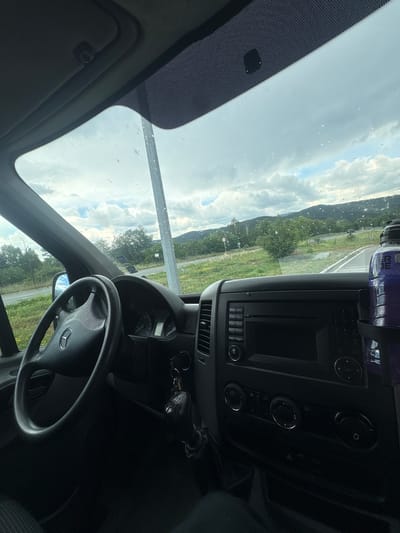 The image depicts the interior of a Mercedes-Benz vehicle, likely a Sprinter van or truck, viewed from the driver's perspective in Pirdop, Bulgaria. The black three-spoke steering wheel, featuring the Mercedes-Benz emblem, dominates the left foreground. To its right, a dark grey dashboard incorporates an instrument cluster, partially obscured, and a central console unit. This console includes a radio/media player with a dark screen and an array of buttons, numbered 1 through 9 on the right side. Below this, three rotary knobs and additional buttons control the HVAC system. A set of vehicle keys with a keyfob hangs from the ignition, positioned between the steering wheel and the central console. A black bottle holder is affixed to the right side of the console, containing a large, purple-tinted plastic bottle, partially legible as "POWERADE".

The windshield is speckled with numerous small water droplets and appears to have some dirt or streaks. A small black rectangular sensor is visible on the upper central portion of the windshield, integrated into the fritted area.

Through the windshield, a cloudy daytime sky with large grey and white clouds is visible. The exterior scene shows a two-lane asphalt road curving to the right, bordered by green grass, bushes, and deciduous trees. In the mid-ground, a tall, light-grey utility pole stands prominently. Distant forested hills or mountains form the backdrop. On the left side of the road, a circular white sign with a red border is partially visible, and further back, another rectangular white sign. A dark-colored vehicle is faintly discernible on the road in the far distance. No individuals are visible within the frame.