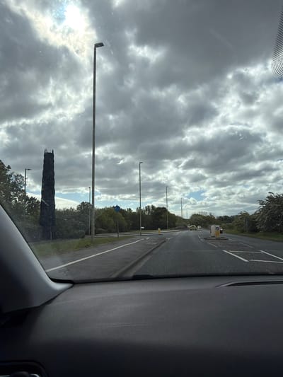 The photograph, taken from inside a vehicle, depicts a view of a roadway and overcast sky in South Tyneside, United Kingdom. On the left of the road, a dark, tower-like sculpture stands. There are several lampposts along the side of the road. The roadway has painted white lane dividers. The sky is filled with a dense pattern of grey and white clouds. The interior of the vehicle is visible on the right side of the image.