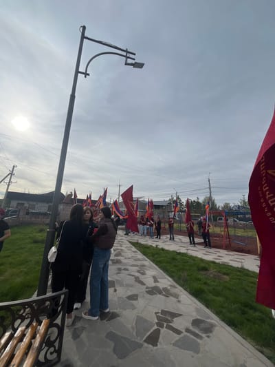 The image shows a scene in the city of Akhuryan, Armenia. A group of people are lined up along a paved walkway, each holding a red flag with Armenian script. Some also hold the Armenian tricolor flag. The walkway is bordered by grass and stone pavement. A lamppost stands near the foreground, and a park with playground equipment is visible in the background. Some buildings can also be seen in the mid-ground. The sky is overcast. A few people are standing or sitting in the foreground on a bench, obscuring part of the view.