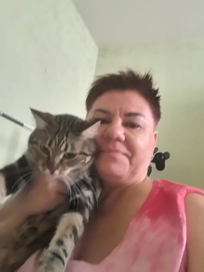 A person with short, spiky, dark red hair holds a tabby cat close to their face. The person is wearing a pink, tie-dye tank top. The cat is looking away from the camera with its ears perked. The background is a light green wall, with what appears to be a black wall decoration to the right of the person's head. The image is taken indoors, suggesting a domestic setting. There is no indication of location in the city of Верхнетемерницкий, Russia.