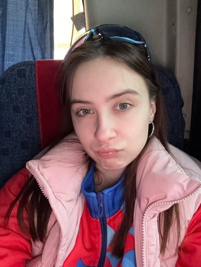 A young woman with fair skin, brown hair, and green eyes is positioned lying down with her head tilted slightly to the left. She is wearing sunglasses perched on her head, a silver earring, and a colorful jacket with pink, red, and blue sections. The background appears to be the interior of a vehicle or airplane, with a seat visible behind her that is patterned with blue and red.
There is no data on this image to support this as being in the city of Челва, Russia.