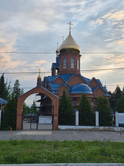 The image shows an Orthodox church under construction in Zelenaya Roshcha 2, Russia. The church is made of red brick with a blue roof and golden domes. The main dome is large and round, topped with a cross, and there are smaller domes as well. Scaffolding is visible around parts of the church, indicating ongoing construction. An arched brick gateway with a black metal fence leads to the church grounds. Several tall, green evergreen trees are planted along the fence. The sky above is cloudy with hints of sunset colors. In the foreground, a street with a crosswalk and two orange traffic cones is visible, separating the viewer from the church property. The overall scene depicts a religious site in a semi-urban or suburban setting, with clear signs of development. No people are visible in the image.