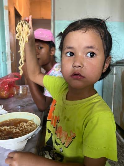 In a dimly lit interior, possibly in Şamaldı-Say, Kyrgyzstan, two young girls are seated at a table. The girl in the foreground, with dark hair and wide, expressive eyes, wears a lime green t-shirt. She holds a fork with curly noodles suspended above it, as if examining them. Her expression is a mixture of curiosity and slight apprehension. Her skin appears somewhat sweaty. Behind her, another girl, wearing a pink baseball cap, is partially visible, also holding noodles. A bowl of ramen noodles sits on the table in front of the foreground girl. Red plastic bags and other food items are also present on the table, which is covered by a marble-patterned surface. The background suggests a simple, possibly domestic setting with plain walls and furniture.