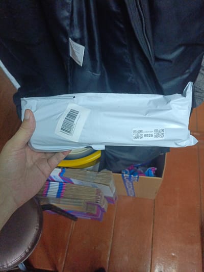The image shows a person's hand holding a white package with a label on it. The package appears to be a small item wrapped in plastic. The background is blurry, but it seems to be an indoor setting with a dark fabric object behind the package and a cardboard box with some items stacked on top in the lower right corner. There is also some sort of yellow circular object visible beneath the package.
The label on the package has a barcode and some text. There are also two QR codes and a number "5926" printed on it. The text on the label is in Cyrillic script and appears to be shipping or product information.
The overall scene suggests someone is holding a recently received package or is about to package something. The lighting is somewhat dim, and the image quality is not very high, making it difficult to discern finer details of the surroundings. However, the context provided places this scene in Ухманы, Russia.