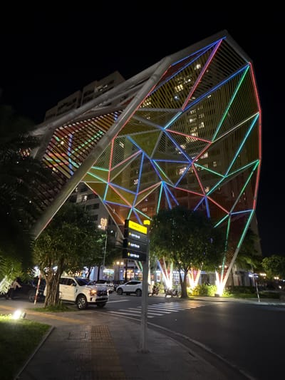 The image depicts a nighttime scene in the city of Dĩ An, Vietnam, specifically featuring a modern building with a distinctive geometric, angular facade illuminated by colorful LED lights. The building's structure appears to be a lattice of metal beams forming triangular and polygonal shapes, outlined with red, blue, green, and yellow lights. Behind this illuminated structure, a taller, multi-story building is visible, with its windows lit from within, suggesting it's occupied.
In the foreground, a street is visible with a pedestrian walkway on the left. Several cars are parked or driving on the street, including a white SUV prominently positioned near the walkway. Two motorcycles are also visible parked on the street. Lush green trees are planted along the street and around the building's base, some of which are brightly lit by ground-level spotlights. A street sign with Vietnamese text and directional arrows is mounted on a pole. The overall impression is a vibrant urban environment at night, characterized by modern architecture and colorful lighting.