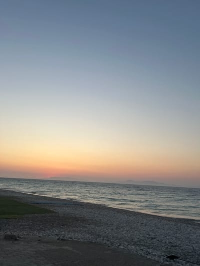 This is a landscape image of a serene beach at dusk. The image captures the gradient of a sunset sky, transitioning from a soft blue to warm hues of orange and yellow, indicating the time of day is either sunrise or sunset. On the right side of the frame, the calm, wavy sea meets a pebbled beach. The pebbles are varied in size and color, creating a textured surface. Further inland, there appears to be a patch of green grass and a paved pathway or promenade, suggesting an accessible coastline. In the far distance, the faint outline of hills or mountains can be seen against the horizon. The overall mood of the image is tranquil and peaceful, typical of a quiet beach scene during twilight. There is no visible text in the image.