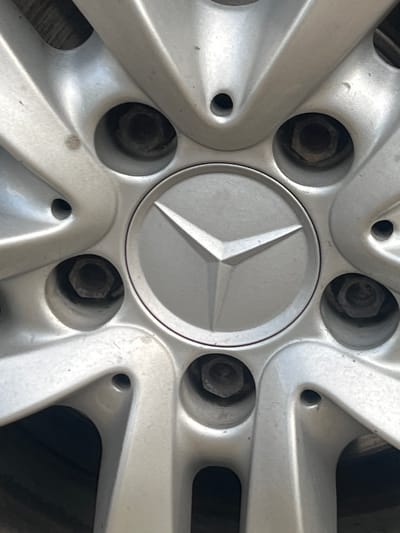 The image shows a close-up of a silver Mercedes-Benz wheel. The central hub features the iconic three-pointed star logo within a circular emblem, outlined by a dark band. Around the hub, there are five visible lug nut holes, each containing a hexagonal lug nut. The wheel has a multi-spoke design, with the spokes radiating outwards from the center. The metal appears to be slightly worn, with some minor scuff marks visible. The lighting suggests it might be daytime, possibly outdoors, as there are shadows and reflections on the metallic surface. There is no text visible in the image, other than the Mercedes-Benz logo. The setting is implied to be Colonia Bod, Romania, based on the provided context. The image focuses solely on the wheel, with no people or other objects clearly depicted.
