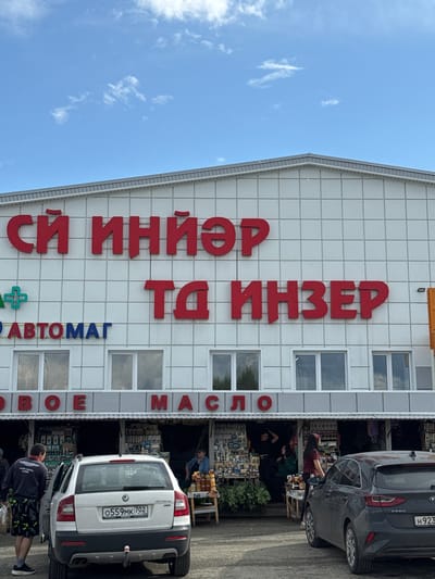 The image shows a street-level view of a commercial building with large red lettering on its facade. The lettering appears to be in Cyrillic and reads "СЙ ИНЙЭР ТА ИНЗЕР", which is the name of the town Inzer, Russia. Below the main signage, there are smaller signs and windows. One sign to the left reads "+ АВТОМАТ" and another, partially visible, reads "СЕВОЕ МАСЛО". The building is white with a grid pattern of darker lines. There are multiple windows across the upper level of the building. The lower level of the building features open market stalls with people behind them selling goods. Two cars are parked in front of the stalls. The car on the left is a white station wagon with a license plate that reads "0559MK 702". The car on the right is a dark grey hatchback. Several people are visible, some interacting with the stalls and others standing near the cars. The environment appears to be an outdoor market on a cloudy day.