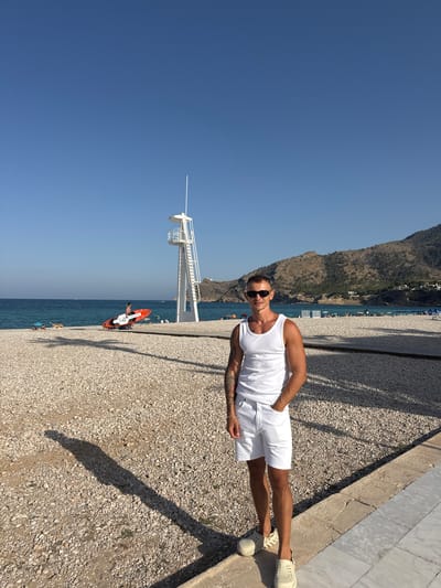 This image depicts a man standing on a pebble beach in L'Alfàs del Pi, Spain. He is wearing a white tank top and white shorts, with sunglasses on. The man has tattoos on his left arm and is standing with his right hand in his pocket. Behind him, the turquoise sea is visible, with a lifeguard tower on the right side of the frame. Further in the distance, mountains with sparse vegetation form the backdrop. The beach is sparsely populated with a few beach umbrellas and people scattered along the shore. The sun is shining, casting shadows on the ground. The overall scene conveys a sense of a relaxed summer day at the beach.