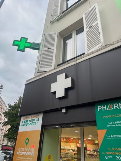 The image depicts the exterior of a pharmacy located in Levallois-Perret, France, on an overcast day. The pharmacy occupies the ground floor of a light-colored, multi-story building featuring windows with open, white louvered shutters on the upper levels.
A prominent, illuminated green LED cross sign extends horizontally from the building's facade, displaying the text "LUCENO" and a phone number "01 47 10 90 20". Directly above the storefront entrance, a large, non-illuminated white cross emblem is affixed to a dark gray/black fascia. This fascia exhibits vertical streaks, possibly from water runoff, and scattered white specks below the white cross.
To the right of the glass entrance, a vertical green and white sign identifies the establishment as a "PHARMACIE," listing "Luc ABITBOL Docteur en Pharmacie" and indicating operating hours from Monday to Friday, 8h30 to 20h. To the left of the entrance, a vertical orange and white banner advertises "VOTRE SANTÉ, C'EST TOUT CE QUI COMPTE!" and "VOTRE FIDÉLITÉ RÉCOMPENSÉE."
The glass entrance reveals a brightly lit interior with shelves stocked with various products. A red poppy-themed wallpaper is visible on the rear wall of the pharmacy's interior. A metallic roller shutter, partially open, is visible above the entrance. In the background to the left, glimpses of other urban buildings, parked vehicles, and green foliage from a tree are visible under a gray sky.