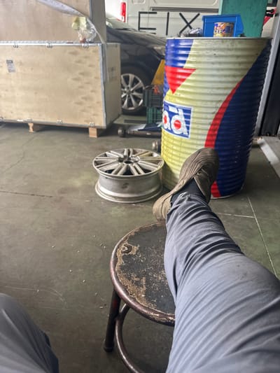 In Marratxí, Spain, a person's right leg and foot, clad in gray trousers and a brown shoe, are prominently featured, resting on a stool in a workshop or storage area. The stool itself is aged, with a worn, dark circular seat and metal legs. To the left, a car wheel rim lies on the concrete floor. Behind the stool, a large, cylindrical barrel with red, yellow, and blue branding stands upright. In the background, a large wooden crate is visible, partially open, revealing a dark vehicle inside. The environment suggests an industrial or automotive setting with a concrete floor and various stored items.