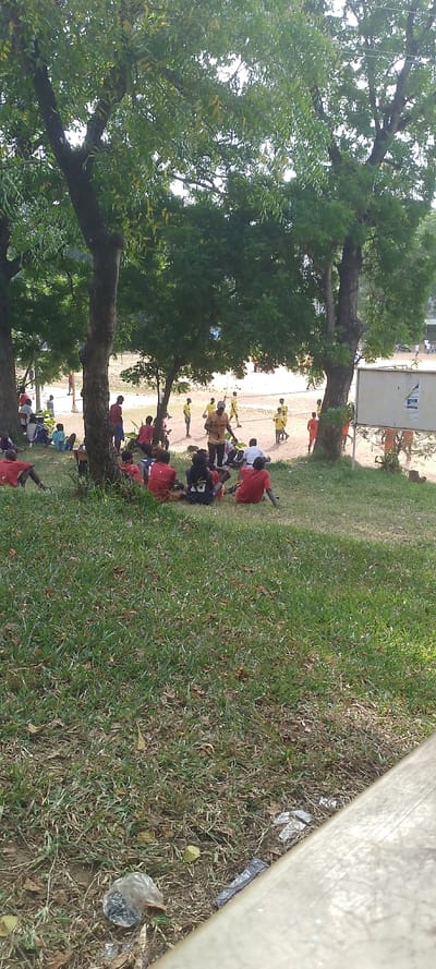 The image displays an outdoor scene in Tudor ward, Kenya, characterized by natural elements and human activity. The foreground consists of green grass interspersed with dried leaves and scattered litter, including a clear plastic bag and a yellow cylindrical object. A light-colored concrete or stone barrier extends diagonally across the bottom right.
In the midground, several large trees with dark, textured trunks and dense canopies of green foliage provide shade. Beneath these trees, approximately 10-15 individuals, primarily young males, are seated or reclining on the grass. Most wear red t-shirts, while a few are in dark-colored tops, suggesting shared group attire. They appear to be resting or observing.
The background reveals a large, open field with light-brown soil or sand. On this field, approximately 15-20 individuals, predominantly young males, are actively engaged. A significant portion of these individuals wear yellow t-shirts, while others are in red t-shirts. One individual in a yellow t-shirt in the central background appears to be addressing or interacting with other players, possibly holding a spherical object. A net structure, consistent with a volleyball court, is visible on the far left of the field. To the right of the field, a white rectangular signboard, exhibiting damage to its lower right section, is affixed to a tree. The scene suggests a sports or recreational gathering in progress or during a break.