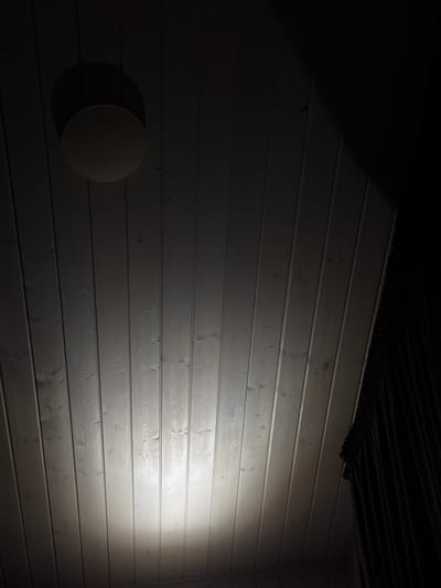 A circular ceiling light fixture is mounted on a wooden planked ceiling. The light from the fixture illuminates a portion of the ceiling, creating a bright, circular glow. The rest of the ceiling is in shadow. A dark curtain is visible on the right side of the image, hanging from the ceiling. The image is taken from a low angle, looking up at the ceiling. The location is Doma 1096 Km, Russia.